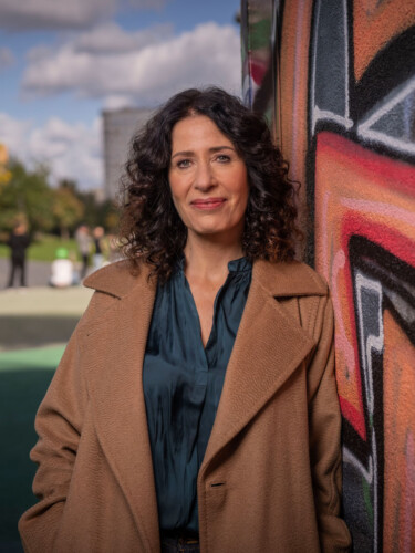 Bettina Jarasch steht an eine mit Graffiti besprühte Wand gelehnt und schaut in die Kamera. Im Hintergrund ein Park, verschwommen sind herbstliche Bäume und Pflanzen zu sehen.